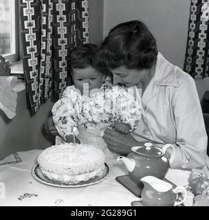 Anni '60, storica, festa di compleanno, seduta ad un tavolo che tiene la sua figlia infantile, una madre che mostra la sua torta di compleanno al bambino, la torta ha candela solitaria, per un anno, Inghilterra, Regno Unito. Foto Stock