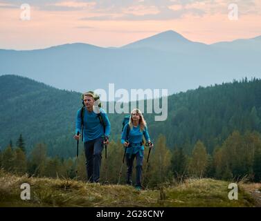 Due persistenti escursionisti professionisti con bastoni da trekking e zaini che si muovono su colline di montagna sullo sfondo di una foresta verde, beskids montagna e cielo viola guardando avanti. Foto Stock