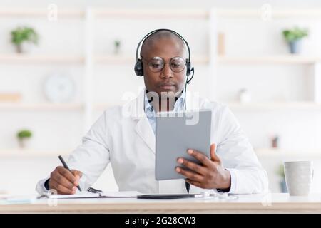 Consultazione online. Medico afroamericano concentrato in cuffie prendere appunti mentre si effettua una videochiamata su tablet Foto Stock