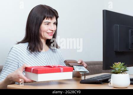 Donna caucasica sorridente seduta di fronte a un monitor che tiene in mano contanti e una scatola regalo rossa. Concetto di shopping online a casa. Acquisto di regali per le feste Foto Stock