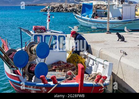 Grecia. Isola di Koufonisi, Cicladi, 23 maggio 2021. Scena di pesca afetr al molo del porto. Pescatore lavorando con le reti su una barca ormeggiata, gabbiani guardare Foto Stock