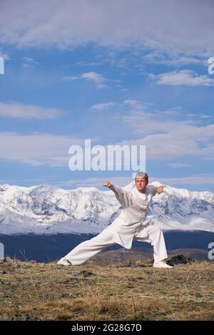 Wushu master in una formazione uniforme sportiva bianca sulla collina. Il campione di Kungfu allena le arti maritiche nella natura sullo sfondo delle montagne innevate. Foto Stock