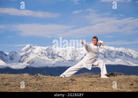 Wushu master in una formazione uniforme sportiva bianca sulla collina. Il campione di Kungfu allena le arti maritiche nella natura sullo sfondo delle montagne innevate. Foto Stock