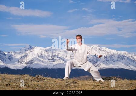 Wushu master in una formazione uniforme sportiva bianca sulla collina. Il campione di Kungfu allena le arti maritiche nella natura sullo sfondo delle montagne innevate. Foto Stock