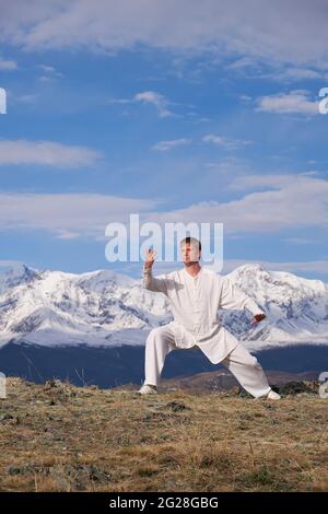 Wushu master in una formazione uniforme sportiva bianca sulla collina. Il campione di Kungfu allena le arti maritiche nella natura sullo sfondo delle montagne innevate. Foto Stock
