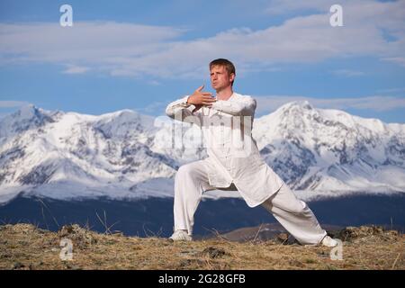 Wushu master in una formazione uniforme sportiva bianca sulla collina. Il campione di Kungfu allena le arti maritiche nella natura sullo sfondo delle montagne innevate. Foto Stock