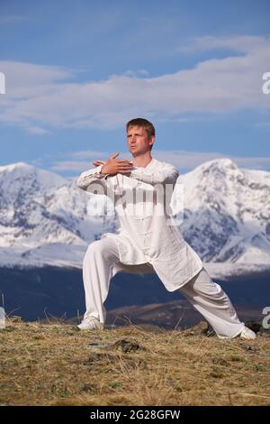 Wushu master in una formazione uniforme sportiva bianca sulla collina. Il campione di Kungfu allena le arti maritiche nella natura sullo sfondo delle montagne innevate. Foto Stock