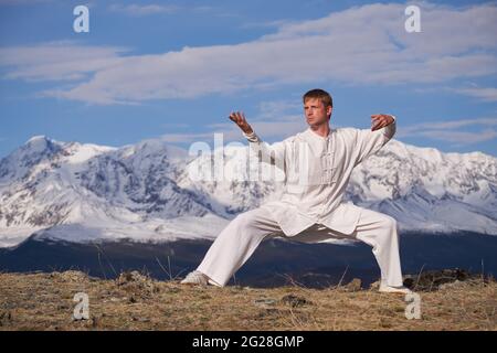 Wushu master in una formazione uniforme sportiva bianca sulla collina. Il campione di Kungfu allena le arti maritiche nella natura sullo sfondo delle montagne innevate. Foto Stock