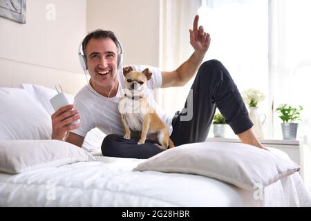Uomo che ascolta la musica dal cellulare con le cuffie ridendo con il suo cane chihuahua a letto con finestra sullo sfondo che entra nella luce del mattino. Foto Stock