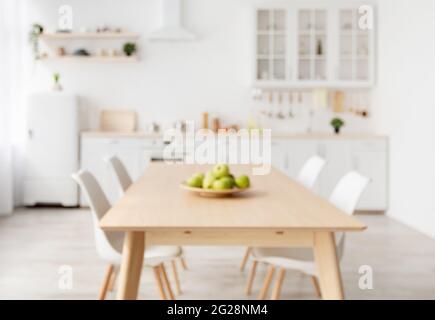 Sfondo sfocato con cucina moderna e luminosa, tavolo da pranzo in legno e mobili bianchi, sedie, utensili da cucina e utensili Foto Stock