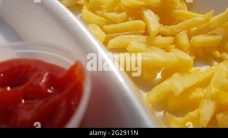 Un primo piano di patatine fritte dorate di colore giallo arancio con ketchup di pomodoro servite in un contenitore di schiuma. Fast food da asporto. Cibo fresco indesiderato. Foto Stock