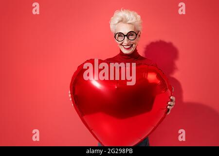 La donna felice tiene il palloncino rosso a forma di cuore. Foto di una donna anziana sorridente innamorato su sfondo rosso. San Valentino Foto Stock