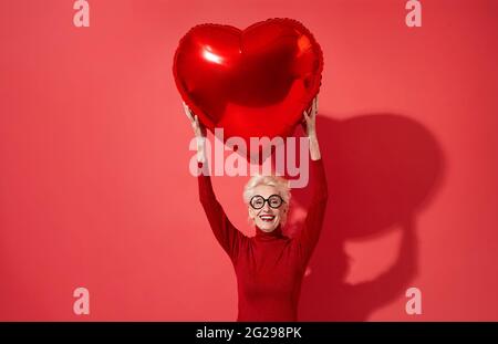 La donna felice tiene il palloncino rosso di forma del cuore sopra la sua testa. Foto di una donna anziana sorridente innamorato su sfondo rosso. San Valentino Foto Stock