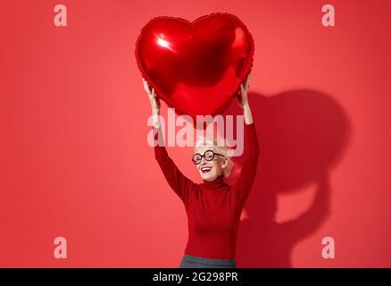 La donna felice tiene il palloncino rosso di forma del cuore sopra la sua testa. Foto di una donna anziana sorridente innamorato su sfondo rosso. San Valentino Foto Stock