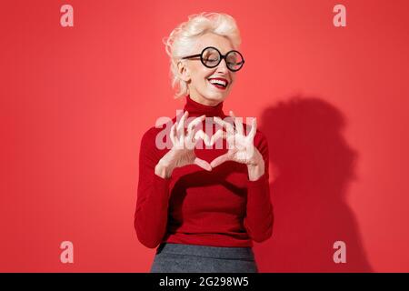 Bella donna mostra il simbolo del cuore, forma segno d'amore con le mani. Foto di una donna anziana sorridente innamorato su sfondo rosso. Sia il mio Valentine Foto Stock