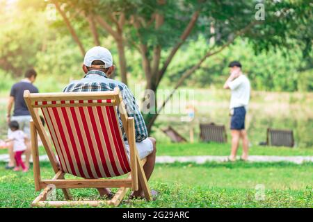 Vista posteriore dell'uomo anziano con cappello bianco seduto sulla sedia da giardino e vicino al tavolo in giardino. Vacanza estiva in un ambiente verde. Persona felice all'aperto Foto Stock