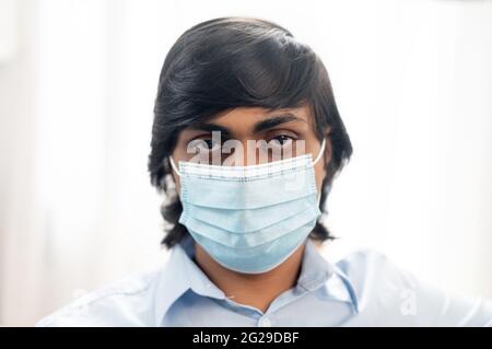Primo piano ritratto di un giovane indiano serio indossando maschera medica chirurgica e una camicia casual elegante blu, l'uomo orientale si protegge da malattie virali, prevenendo la diffusione del virus corona Foto Stock