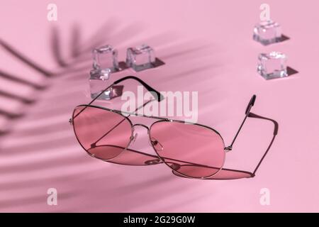 occhiali da sole rosa e cubetti di ghiaccio sotto una palma su uno sfondo pastello. ombre dure dal sole a mezzogiorno. concetto di svago, viaggio e intrattenimento Foto Stock