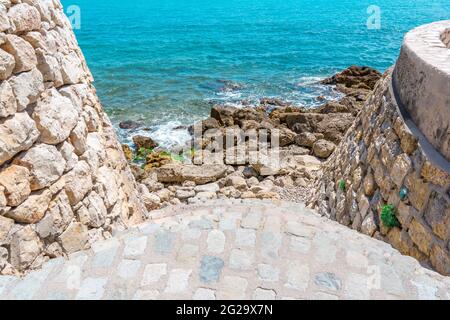 Fortezza e una scala in pietra, che va in mare. Molte grandi e piccole rocce e onde di mare che colpiscono le rocce. Blu mare onde sfondo e cielo bellissimo paesaggio. Foto di alta qualità Foto Stock