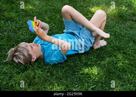 Bambino a piedi nudi sdraiato sull'erba, gioca con il fidget sensoriale pop it giocattolo. Il giocattolo flessibile a bolle d'aria si spinge nelle mani del ragazzo, giocando all'aperto durante il giorno di sole. Foto Stock