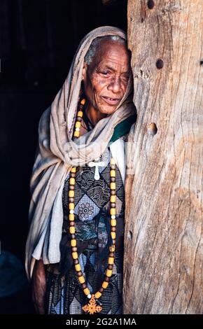 Donna rugosa anziana che indossa collana culturale con mantello appoggiato sul legno in luce del giorno, Etiopia Foto Stock