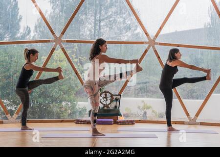 Vista laterale delle donne equilibrate e flessibili che sollevano la gamba e tengono i piedi con le mani sul tappetino in classe per lo yoga Foto Stock