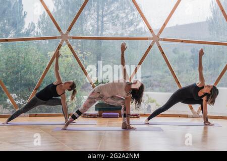 Vista laterale di donne calme equilibrate concentrate su stretching e alzando le mani in su nella postura del guerriero classe rotonda per yoga Foto Stock