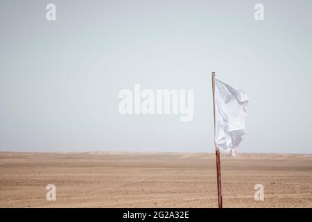 Bandiera bianca appesa al palo contro il deserto sabbioso del Sahara e il cielo grigio senza nuvole Foto Stock