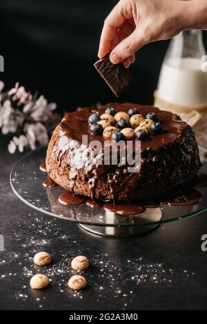 Crop Unriconoscibile Woman utilizzando biscotto al cioccolato per decorare torta fatta in casa ricoperta di ganache al cioccolato e bacche Foto Stock