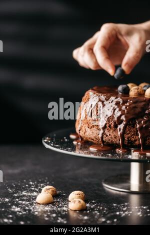 Crop Unriconoscibile Donna utilizzando mirtillo per decorare torta fatta in casa coperto di ganache al cioccolato e bacche Foto Stock