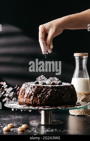 Crop persona anonima spruzzando polvere di zucchero su torta di cioccolato casereccia yummy decorata con sciroppo di cioccolato e dolci assortiti posti su strada di vetro Foto Stock