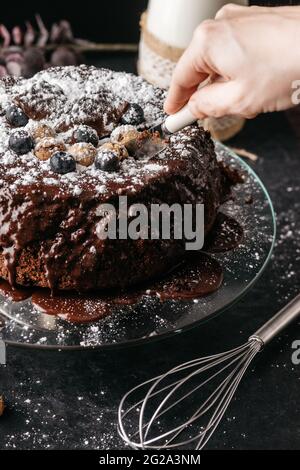 Crop persona anonima tagliando pezzo di torta al cioccolato yummy con coltello posto su piastra di vetro rotonda sul tavolo con frusta e bottiglia di latte Foto Stock