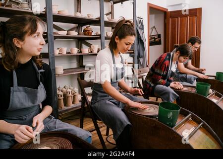 Persone entusiaste in grembiuli che lavorano con la scultura in argilla su ruota in ceramica con l'aiuto di artigiani in grande officina leggera Foto Stock