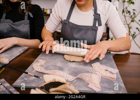 Da sopra la persona di raccolto in grembiule che fa appiattita l'argilla con il perno di rotolamento alla tabella in officina Foto Stock