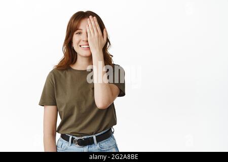 Felice ragazza sorridente coprire metà del viso con la mano, guardare allegro, nascondere un occhio, controllare la vista in negozio eyewear, in piedi su sfondo bianco Foto Stock