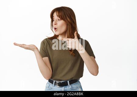 Impressionato giovane donna che punta, guardando la mano aperta, tenendo l'oggetto sul palmo e dire WOW, comprare qualcosa di fresco, consigliare il prodotto, in piedi contro Foto Stock