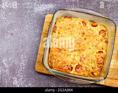 Casseruola con formaggio, cipolle verdi, uova e salsicce in una teglia di vetro su fondo scuro. Vista dall'alto, disposizione piatta Foto Stock