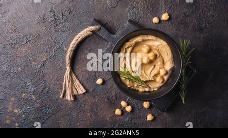 Vista dall'alto del tradizionale spuntino hummus di ceci fatto in casa con rosmarino fresco verde in ciotola nera su fondo di cemento scuro Foto Stock