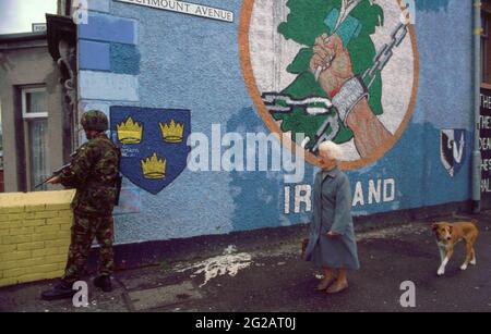 Belfast, Irlanda del Nord. Ottobre 1993. Un soldato dei British Royal Marines in pattuglia su Beechmount Avenue e Falls Road giunzione come una donna anziana e cane passare da un murale repubblicano per un'Irlanda libera. Beechmount Avenue divenne noto come RPG Avenue dopo che una granata a propulsione a razzo fu lanciata dalle forze britanniche da quel luogo. Foto Stock