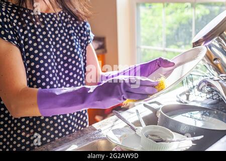 Una donna che indossa guanti da lavoro viola sta lavando i piatti. Sta lavando un piatto di porcellana bianca usando un detergente e una spugna. I guanti proteggono le mani Foto Stock