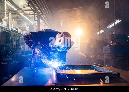 I due handymen che eseguono la saldatura e la molatura sul luogo di lavoro in officina, mentre le scintille "volano" tutto intorno a loro, indossano un lui protettivo Foto Stock