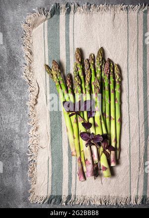 Vista dall'alto di un fascio di asparagi verdi freschi con foglie di basilico viola su una tovaglia dalla linea rustica. Foto Stock