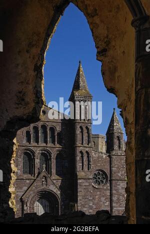 Una finestra gotica rovinata del Palazzo Vescovile del 14th ° secolo accanto alla Cattedrale di San Davide a Pembrokeshire, Galles, Regno Unito, incornicia una torre e parte della cattedrale di fronte ad ovest, Ricostruito in arenaria viola in stili misti e con standard tecnici insoddisfacenti nel 1793 dal celebre architetto neoclassico Regency e Georgiano John Nash, designer di Buckingham Palace, Marble Arch e Regent Street di Londra, e dal Royal Pavilion di Brighton. La facciata doveva essere rimodellata negli anni '1860s, quando iniziò a piegarsi verso l'esterno e necessitò di ulteriori riparazioni importanti negli anni '1990s. Foto Stock