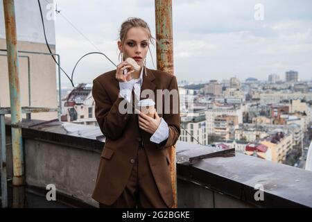 giovane donna in outfit elegante che tiene una tazza di carta e una ciambella sul tetto con paesaggio urbano su sfondo sfocato Foto Stock