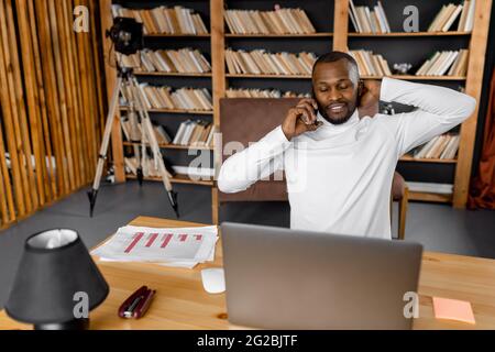 Un dipendente o uno studente di sesso maschile felice sta conversando telefonicamente con un amico o un collega, sorridendo. Gioioso uomo d'affari giovane adulto è seduto al lavoro Foto Stock