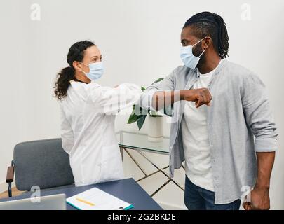 L'uomo afroamericano e il suo medico indossando maschere mediche si salutano l'un l'altro da gomito che urta all'ufficio del medico. Gomito di battuta, battaglia coronavirus epid Foto Stock