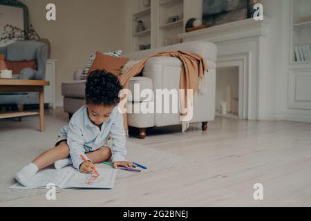 Piccolo ragazzo misto corsa disegno in libro da colorare mentre si passa il tempo libero da solo a casa Foto Stock