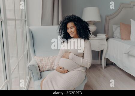 Donna incinta afro americana affascinante guardando in finestra e pensando al futuro bambino Foto Stock