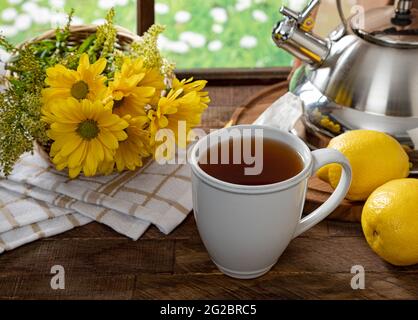 Tazza di tè con limoni e fiori su un tavolo rustico in legno vicino a una finestra Foto Stock