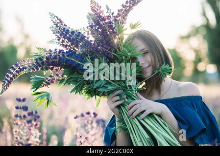 Giovane bella ragazza tiene un grande bouquet di lupini viola in un campo fiorito. Foto estate. Foto Stock